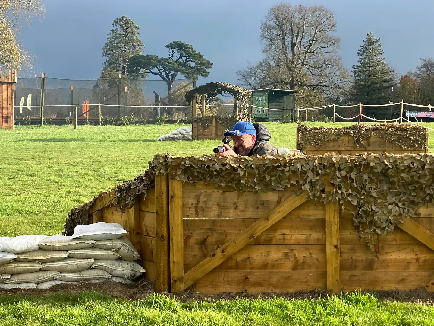 An adult laser tag player in a blue sensor cap aiming over a camouflaged barricade at Escot Park