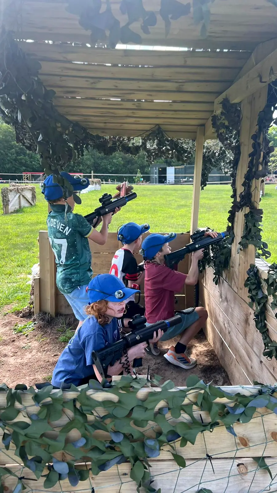 Four children in blue sensor caps playing laser tag from a camouflaged wooden shelter