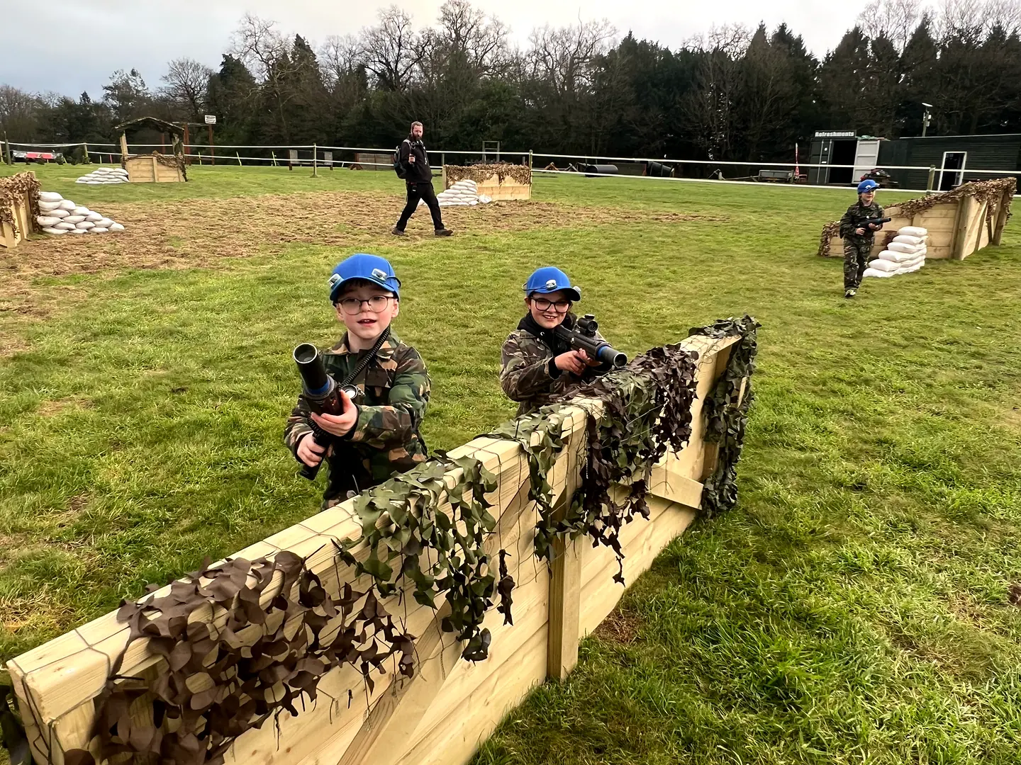 Two children in blue sensor caps playing laser tag behind a camouflaged timber barricade near Exeter