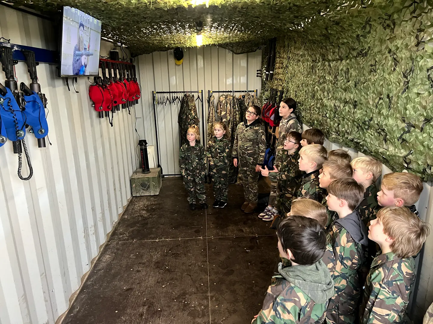Children in camouflage overalls watching the laser tag briefing beside racks of equipment