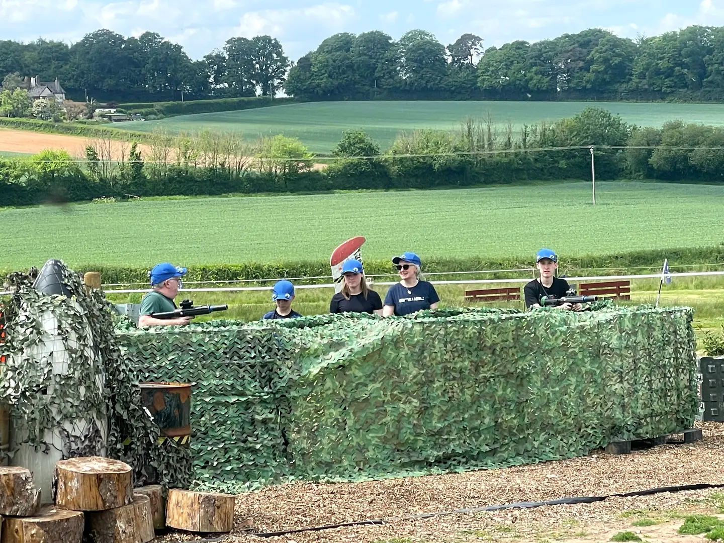 A group of blue-team laser tag players behind a camouflage barrier with Devon fields in the background